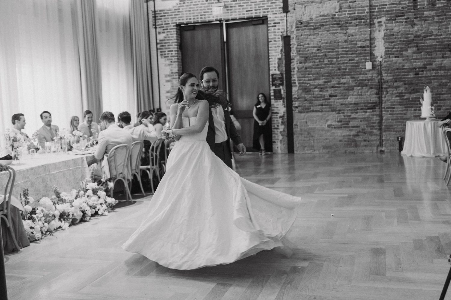 Newlyweds share their first dance in a rustic brick-walled reception venue while guests look on from their seats.
