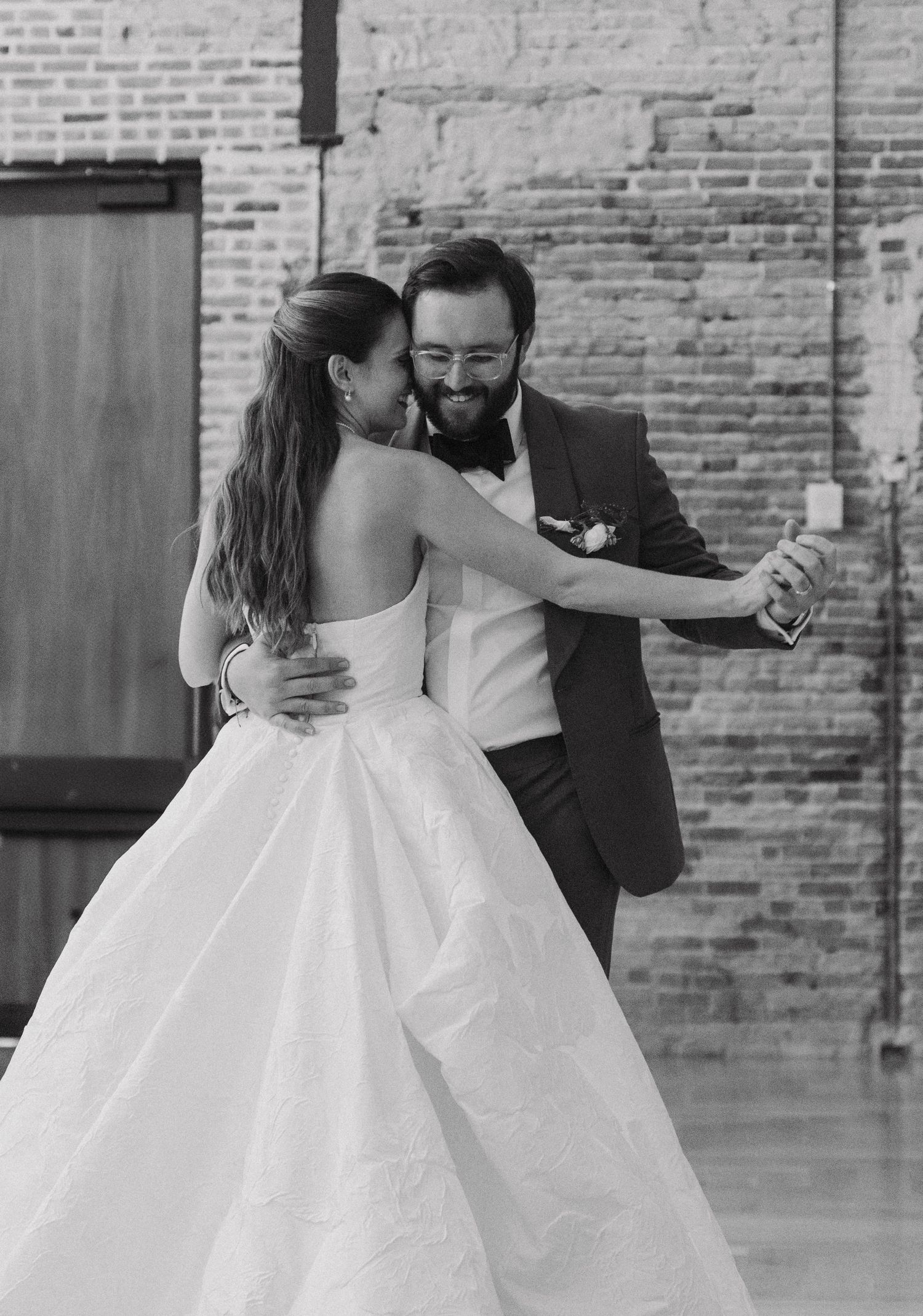 A couple in wedding attire dances together in an intimate sequence of black and white photos against a brick wall.