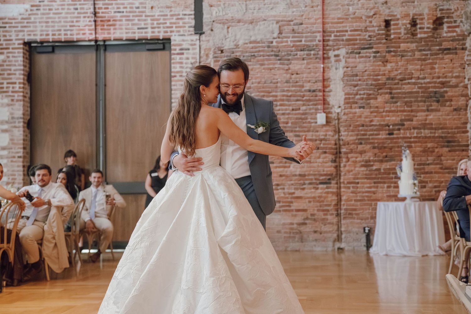 Bride and groom share their first dance in an industrial brick-walled venue with guests watching from tables.