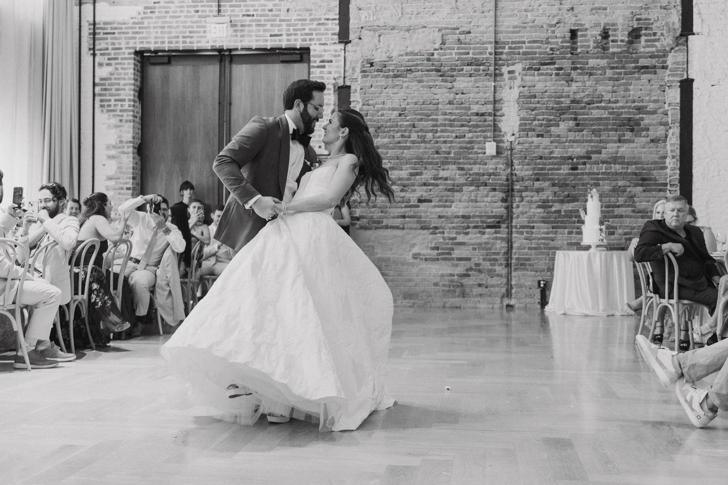 A couple performs their wedding dance in an industrial-style venue with exposed brick walls and seated guests watching.