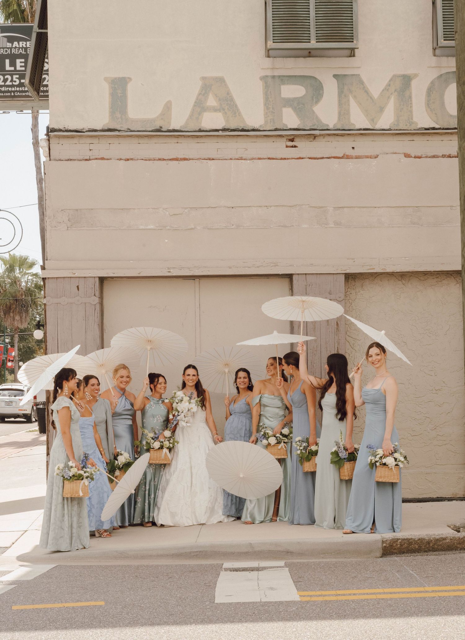 Wedding party in light blue bridesmaid dresses poses against a beige wall outdoors.