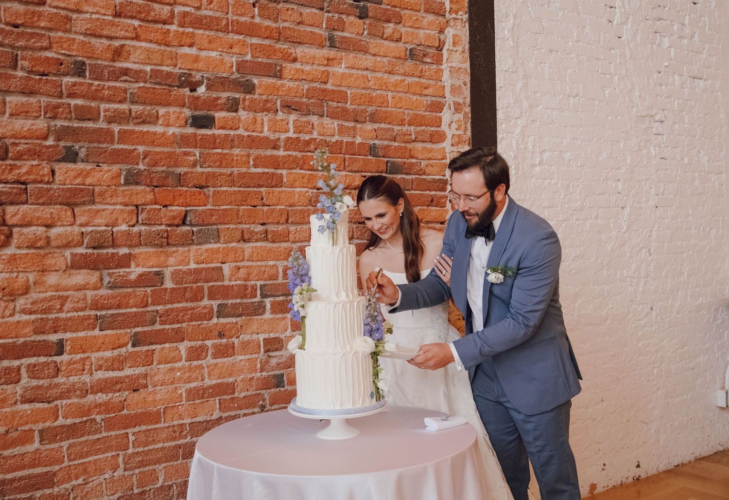 Wedding couple cuts their elegant white tiered cake together against a rustic brick wall backdrop.