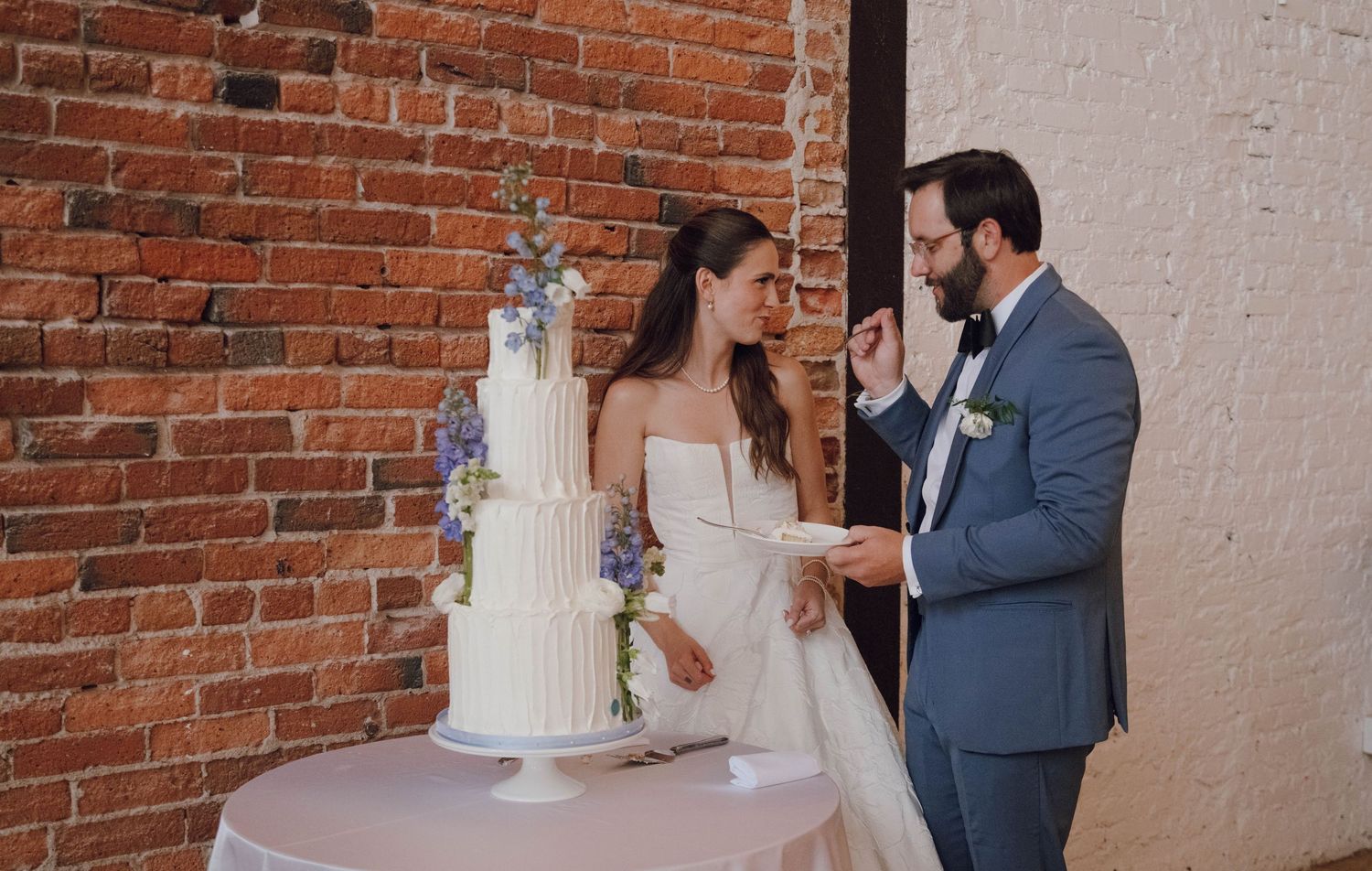 A bride and groom cut into their elegant white tiered wedding cake beside a rustic brick wall.