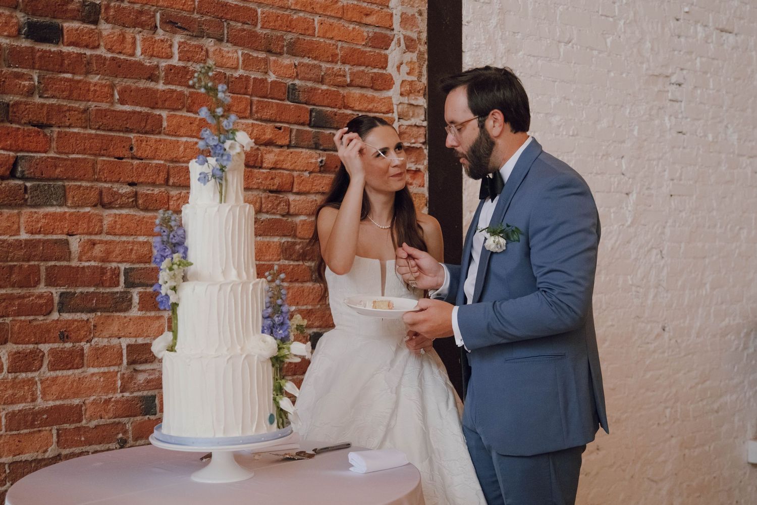 Couple sharing a joyful moment while cutting their elegant white tiered wedding cake against a brick wall.