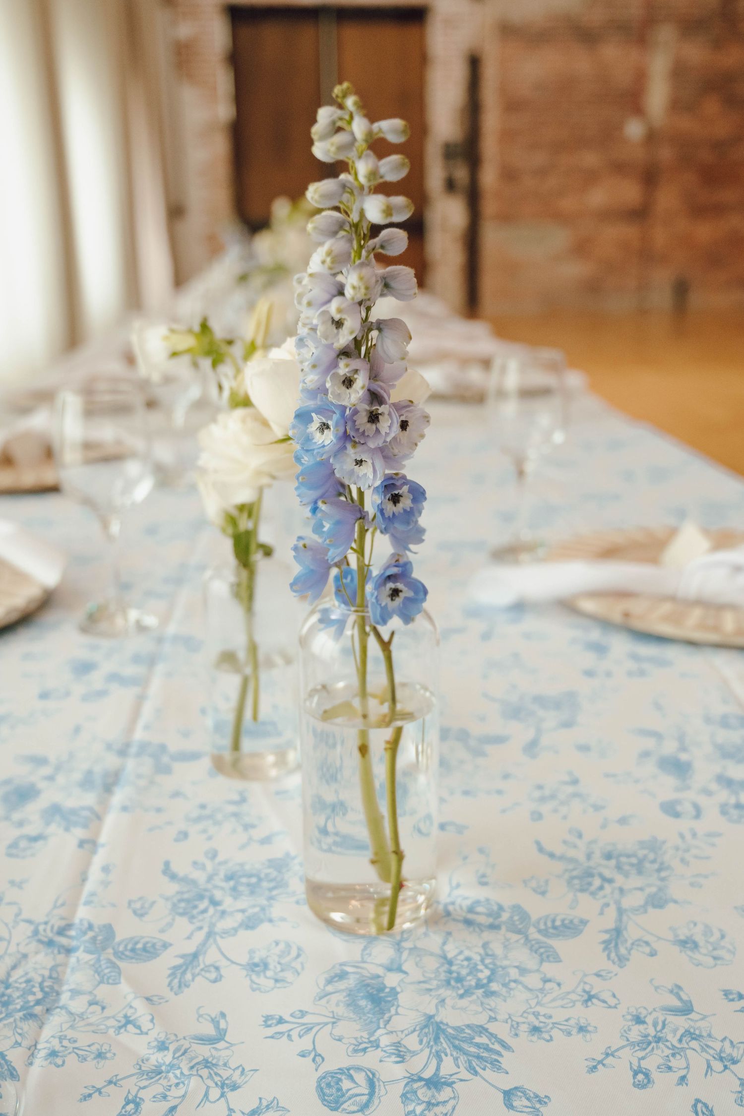 Light blue floral tablecloths with delicate blue delphinium flowers in glass bud vases creating an elegant table setting.