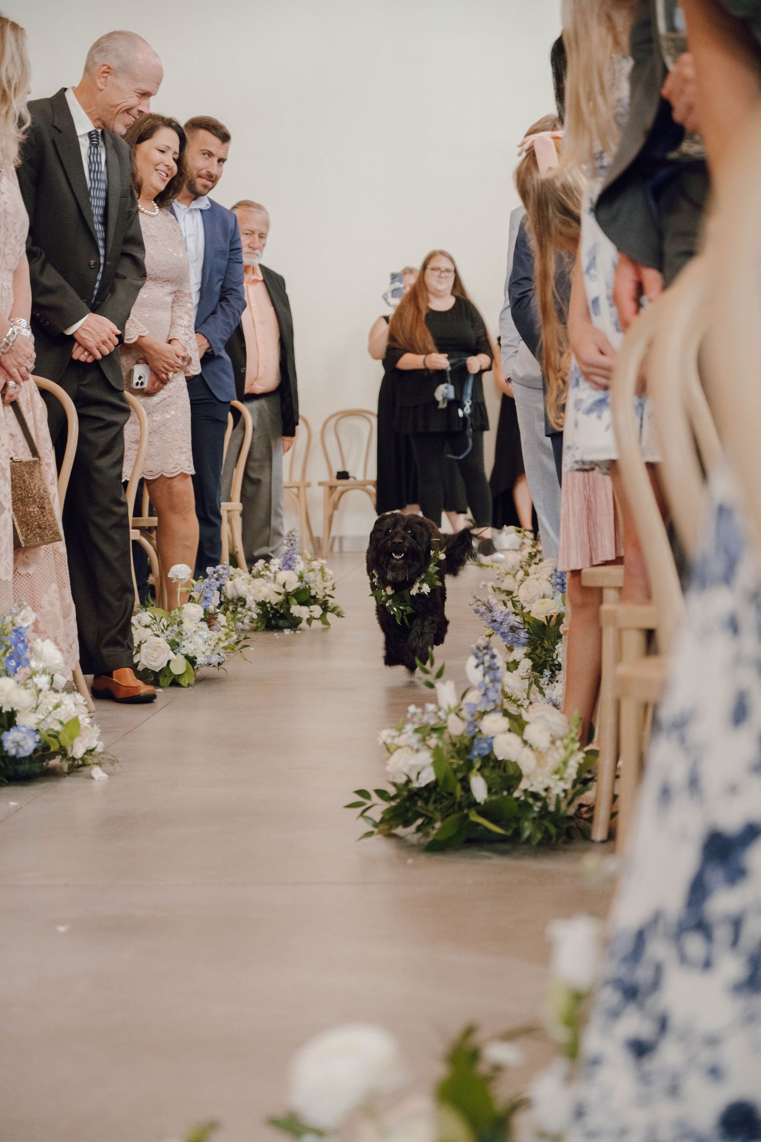 Wedding guests stand in rows with floral aisle decorations on a concrete floor.
