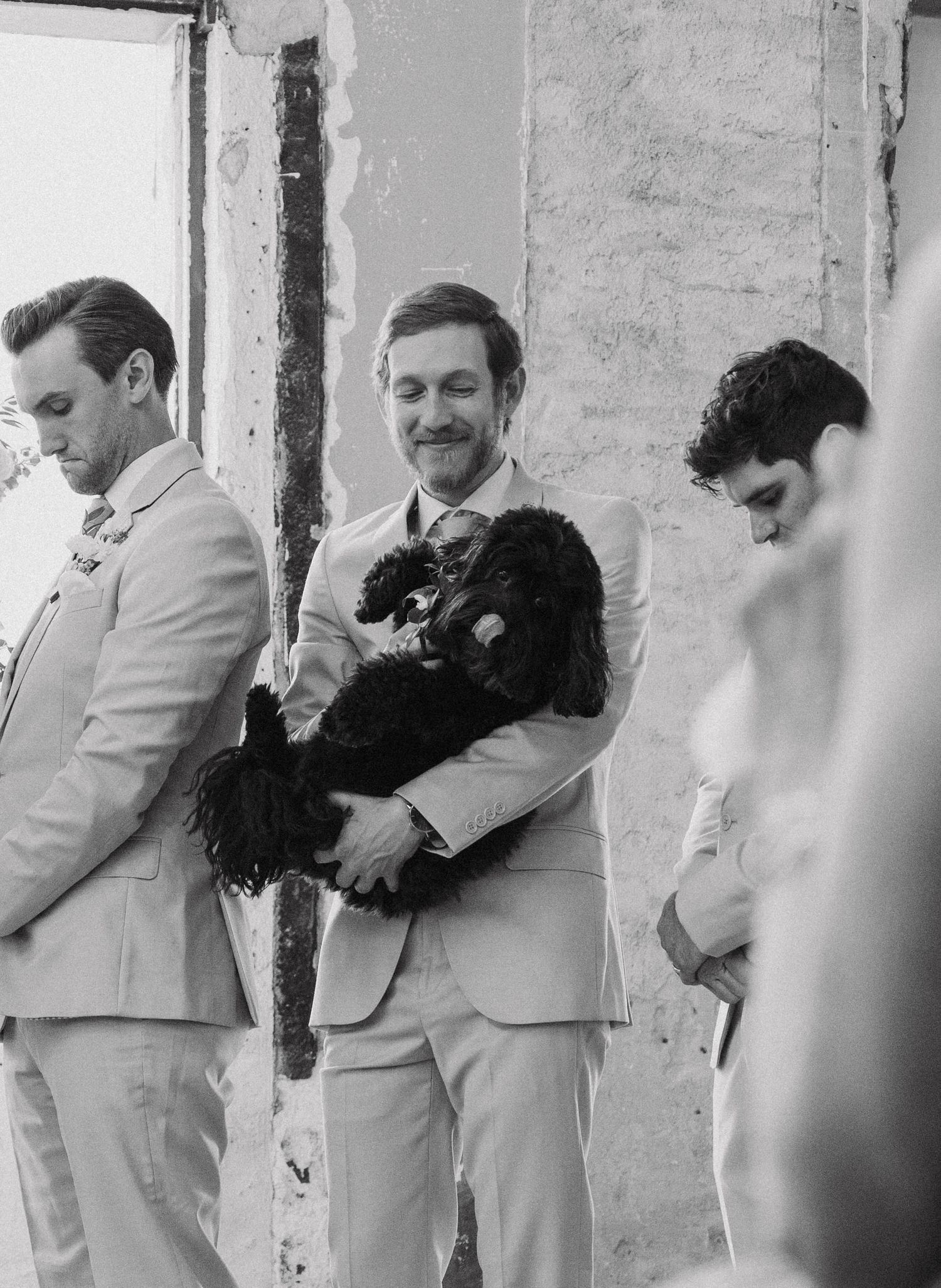 Groomsmen in light colored suits pose against a wall while one holds a black dog.