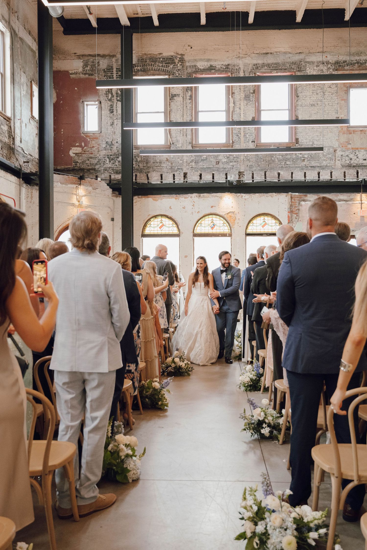 Wedding ceremony in an industrial-style venue with exposed brick walls, wooden chairs and floral aisle decorations.