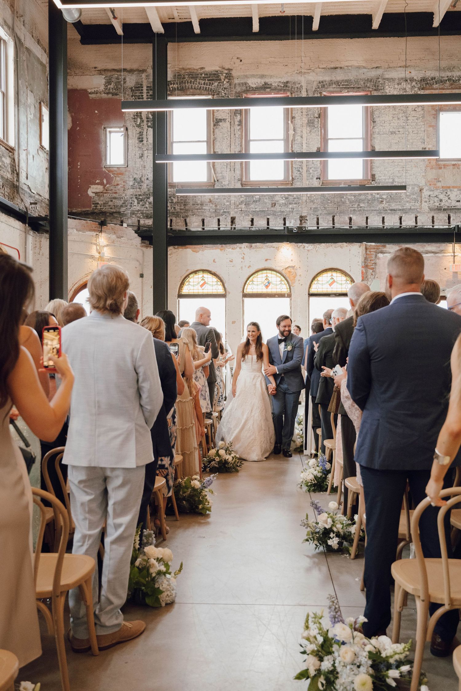 Wedding ceremony in an industrial style venue with exposed brick walls and wooden chairs lined with floral arrangements.