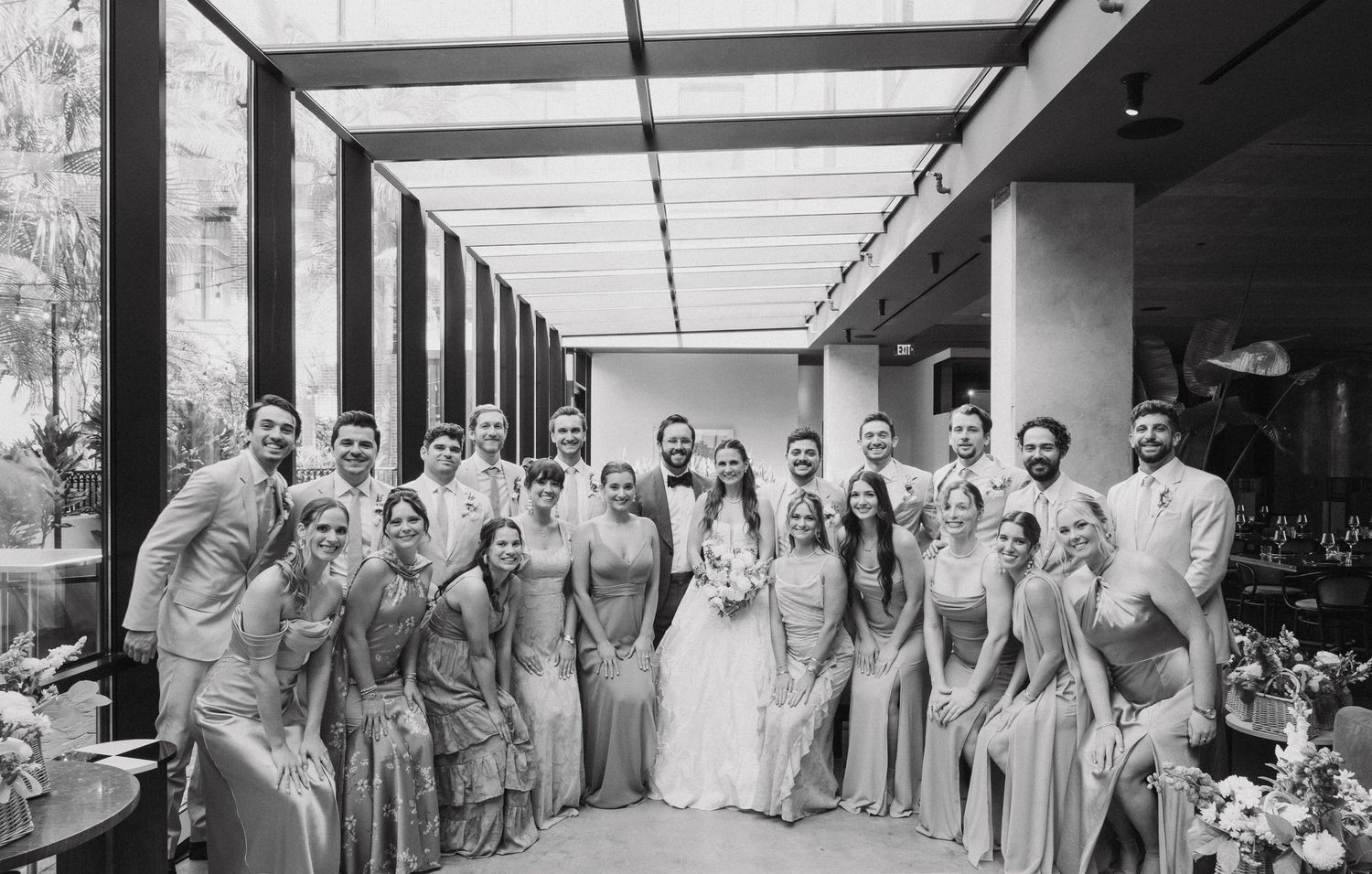 Black and white photo of wedding party members lined up in formal attire at indoor reception venue.