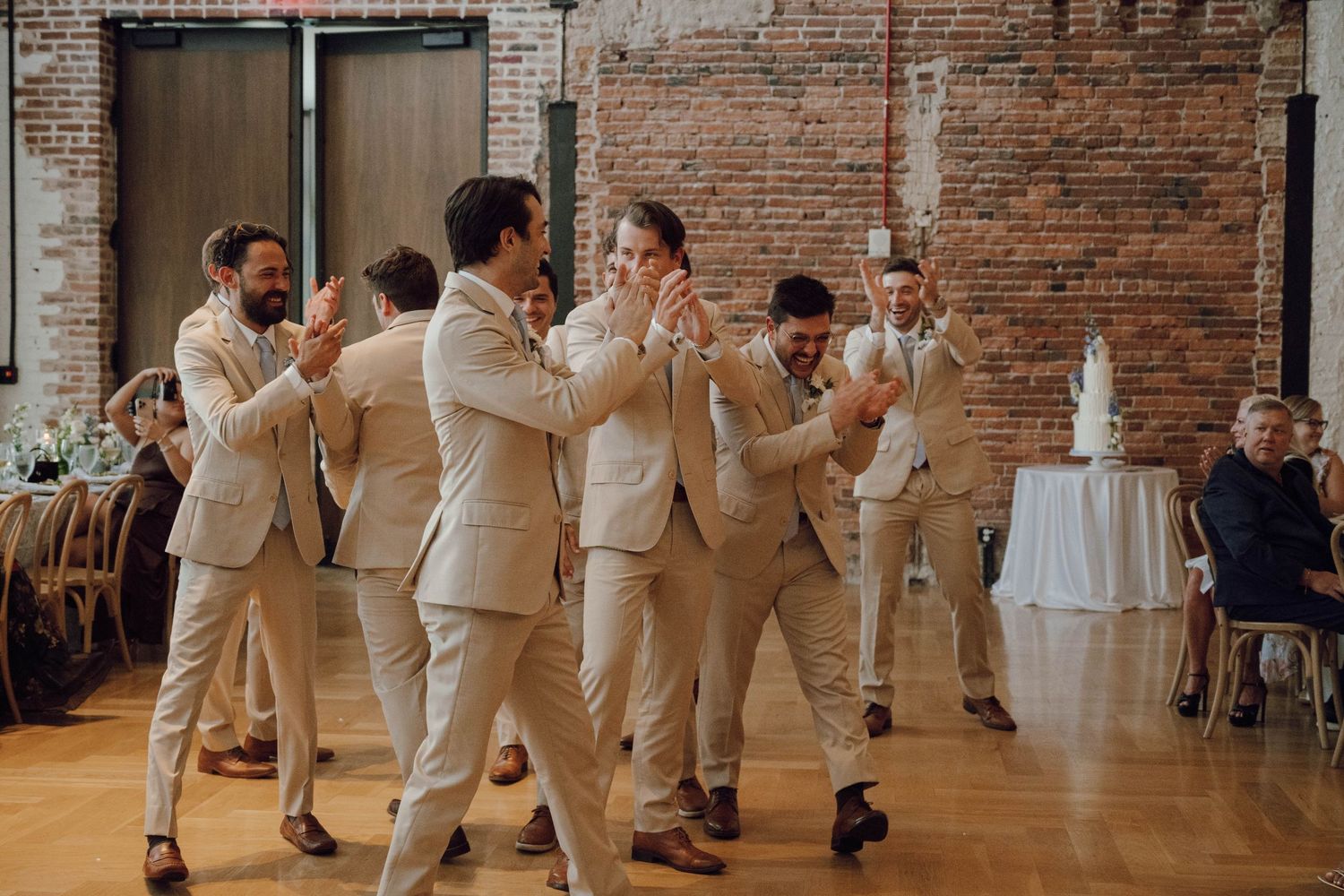 Wedding party in beige suits and white dresses dance together in a rustic brick venue during reception celebration.