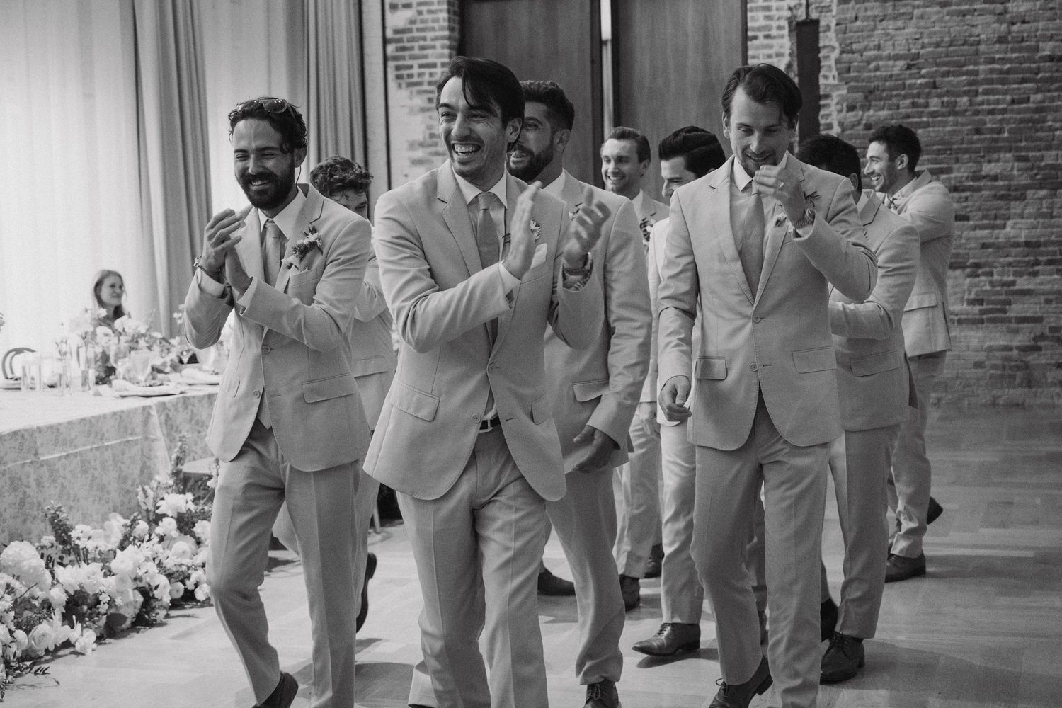 A group of groomsmen in light gray suits walk in line while laughing and smiling during a wedding celebration.