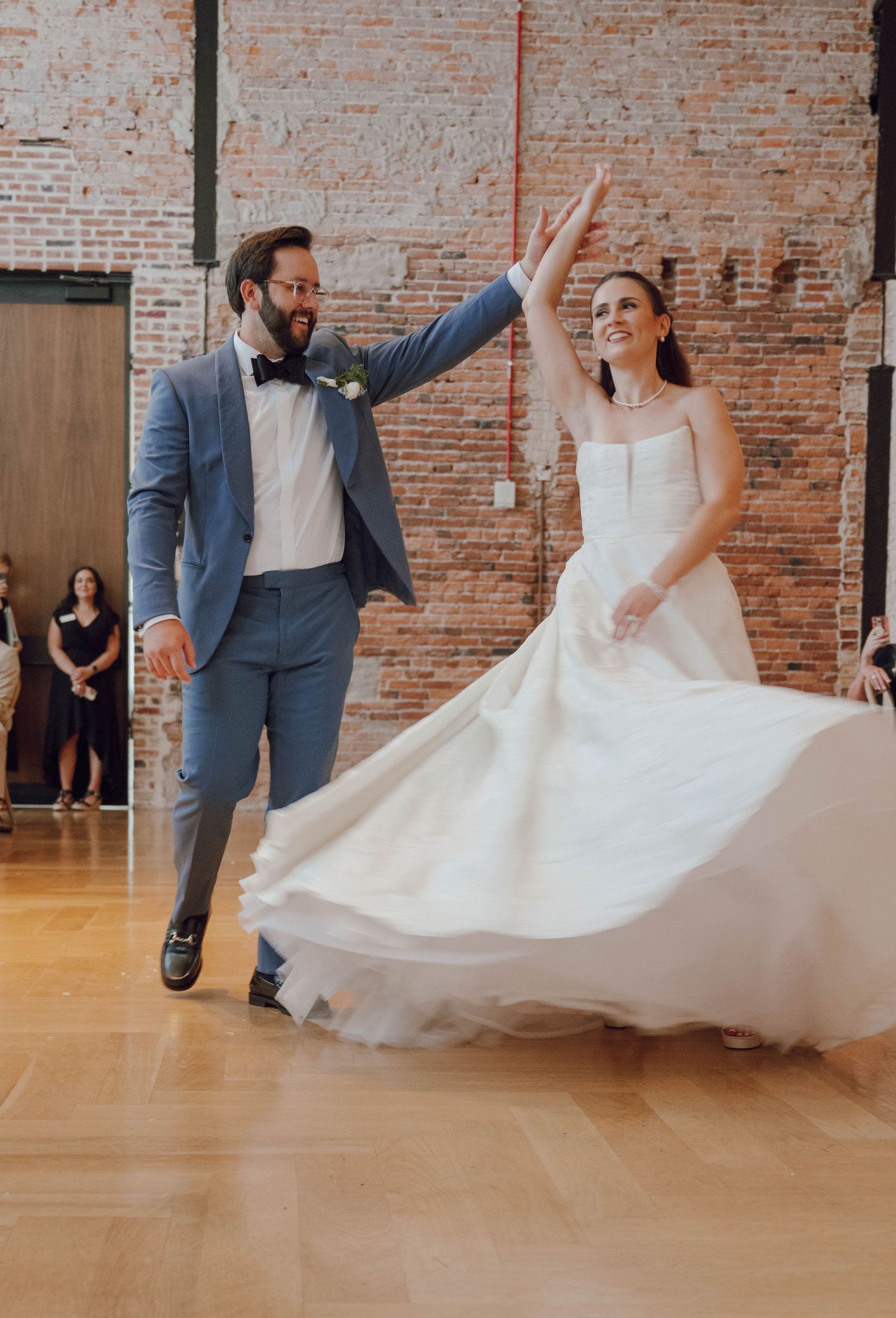 A couple performs a spinning dance move on a wooden floor in front of an exposed brick wall during their wedding celebration.
