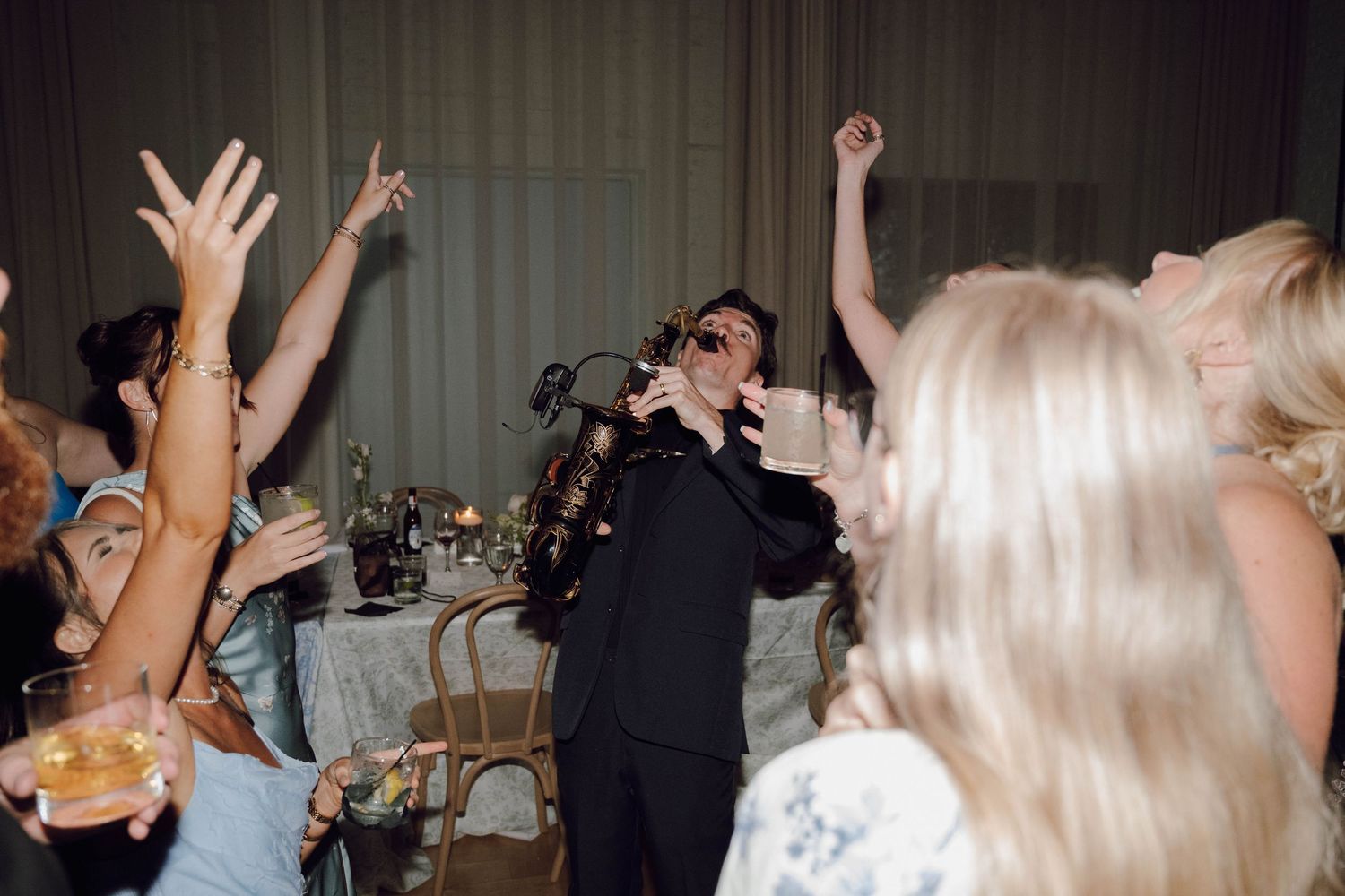 A singer performs with a microphone at a party while people raise their hands and dance in celebration.