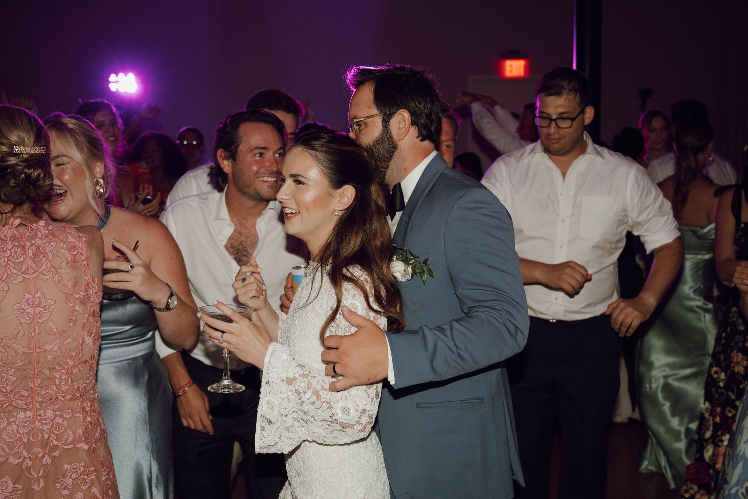 Wedding guests dance and celebrate together on a dimly lit dance floor with purple lighting.