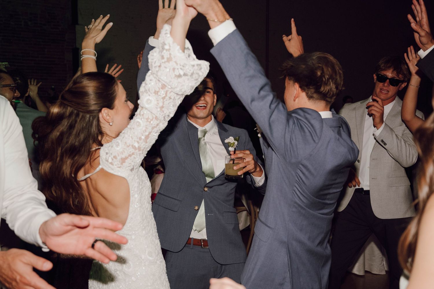 Wedding guests dancing with raised arms on a crowded dance floor during an evening reception celebration.