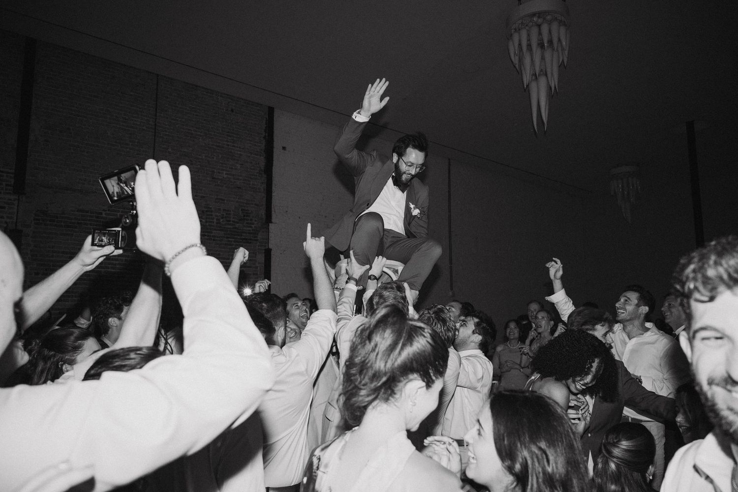 A crowd of people at a lively party raise their hands in celebration on a dark dance floor.