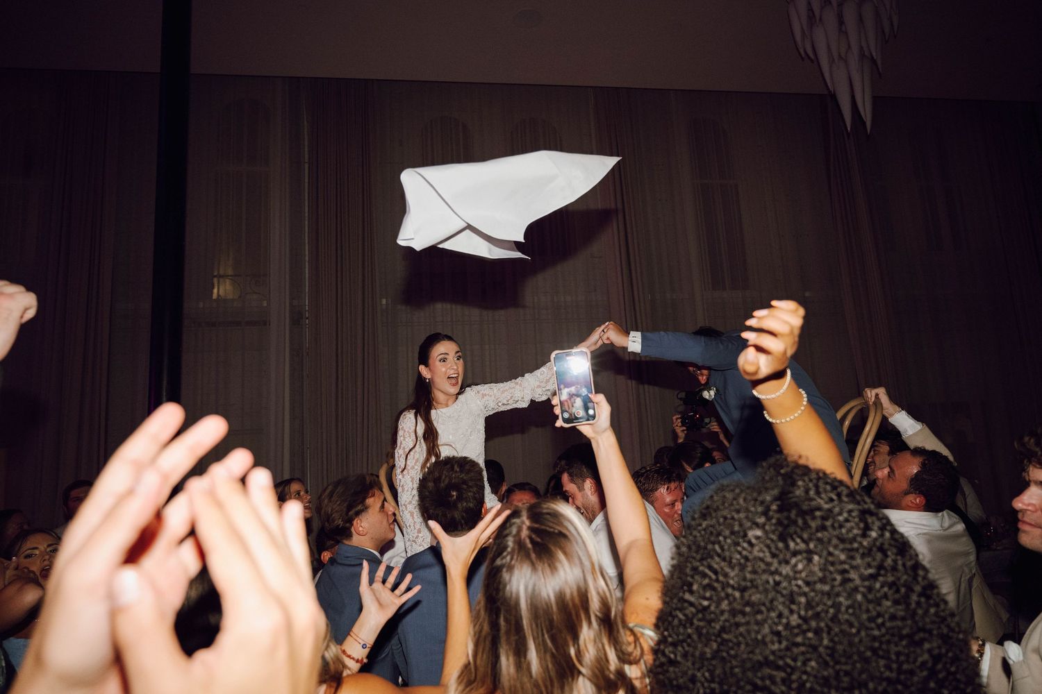 Crowd of wedding guests dancing and celebrating with raised arms on a dark dance floor.