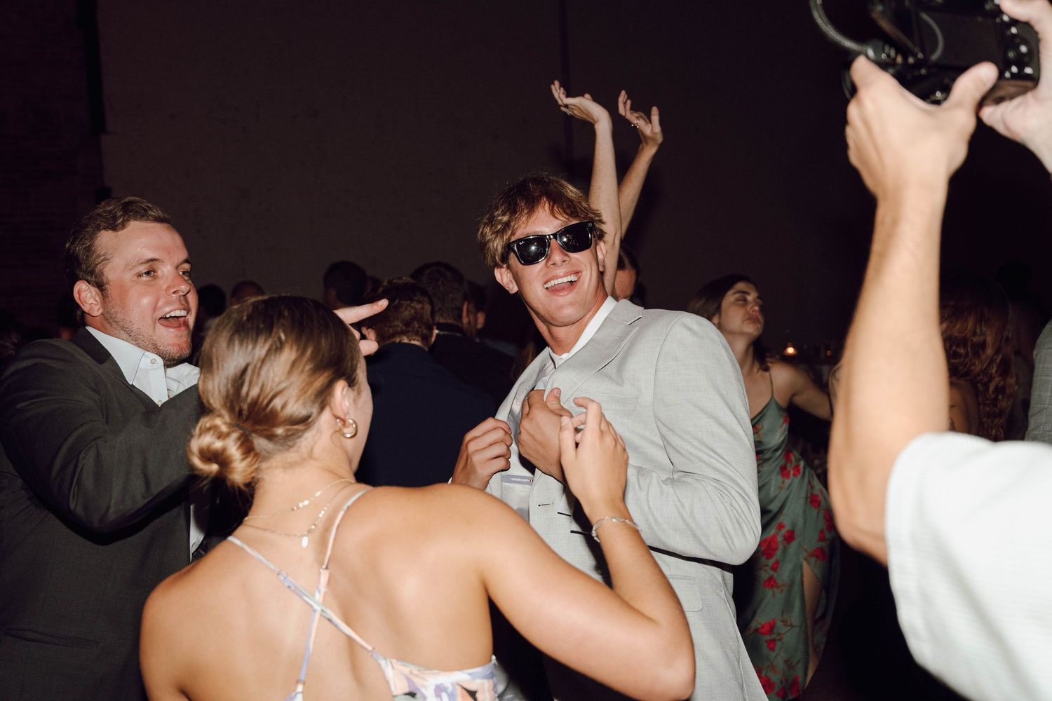 Party guests dancing and celebrating enthusiastically at a dimly lit indoor reception event.