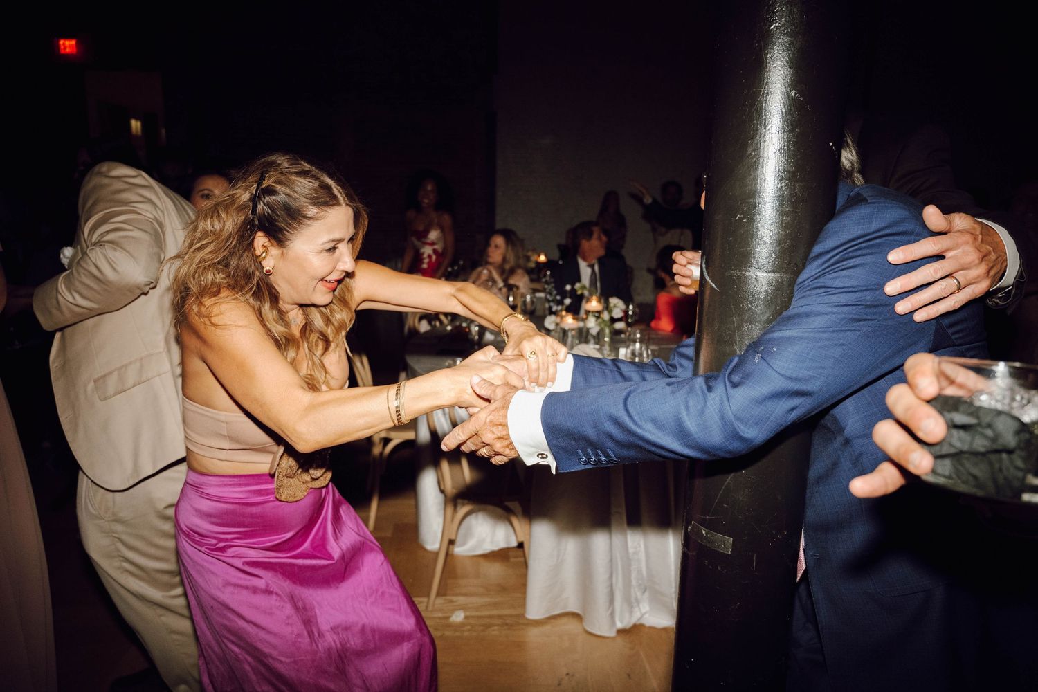 Wedding guests dance energetically on a dimly lit dance floor during an evening reception celebration.