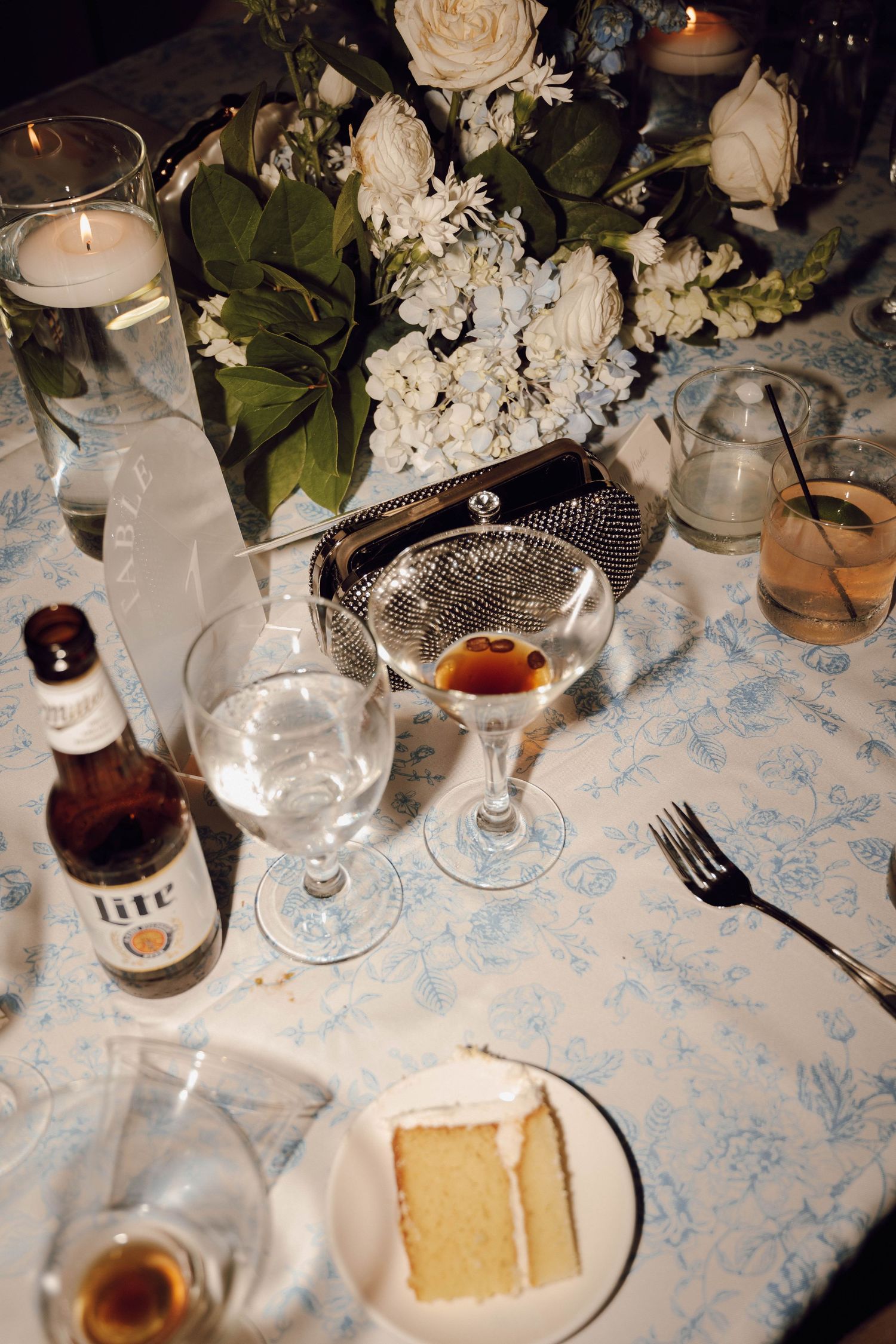 An elegant table setting with cocktails, roses, and a slice of cake on a patterned tablecloth.