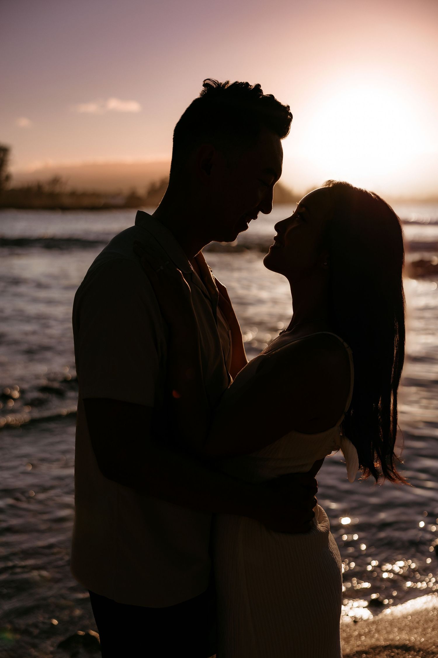 Romantic couple shares intimate moment at sunset by the water's edge, sitting close together on rocky shore.