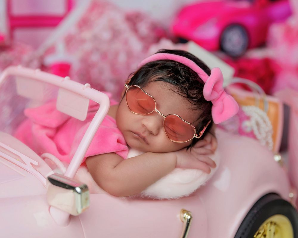 Adorable newborn baby girl sleeping in pink toy car wearing sunglasses and pink bow headband.