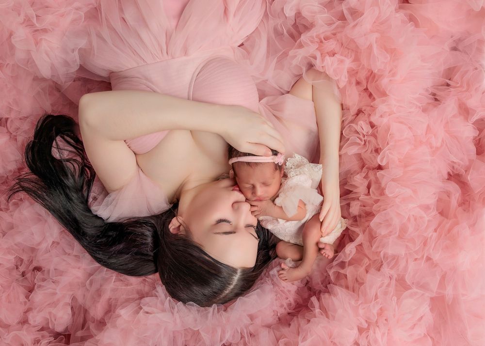Mother in pink tulle dress lying down cuddling her sleeping newborn baby girl.