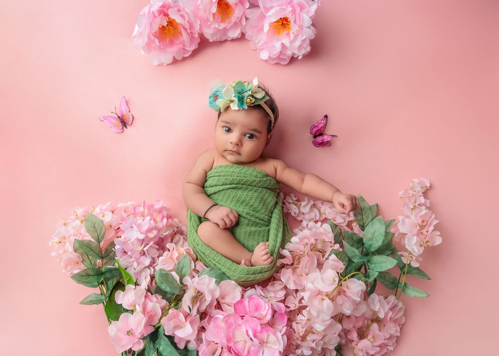 Awake newborn in green wrap lying among pink flowers on pink background.