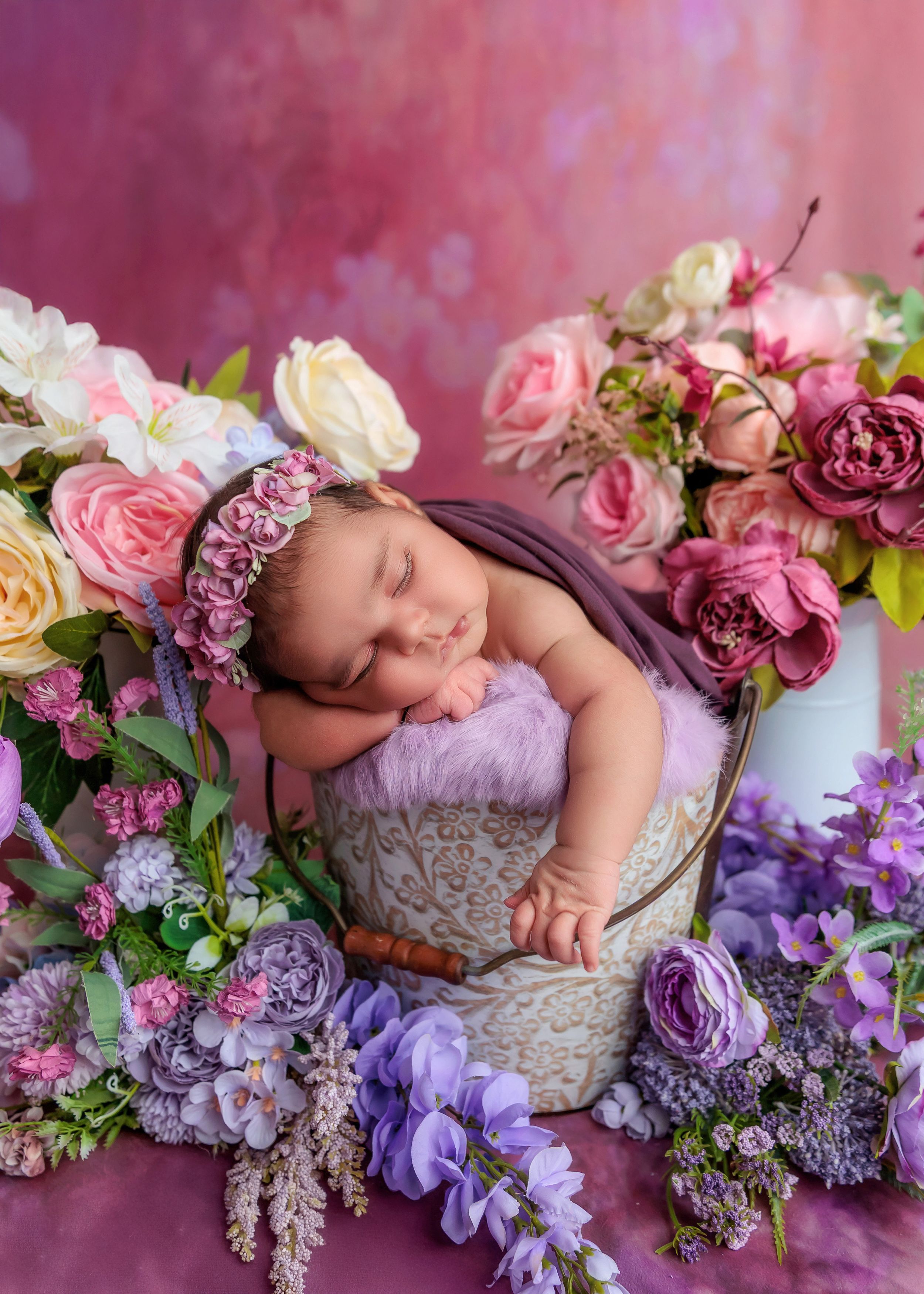 Newborn baby sleeping in bucket surrounded by colorful pink and purple flowers.