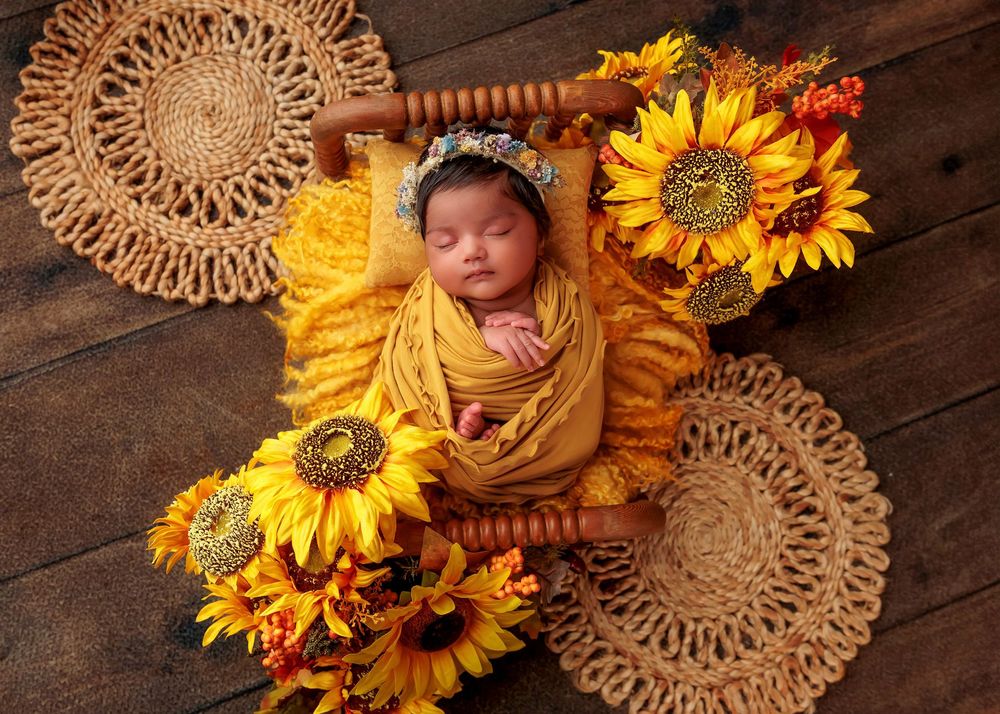 Sleeping newborn wrapped in yellow swaddle with floral crown surrounded by sunflowers on wood floor.