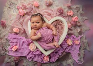 Newborn baby girl in purple wrap posed in white heart frame with pink roses.