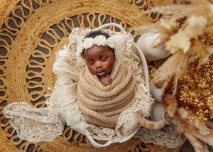 Sleeping newborn wrapped in tan knit swaddle with lace bonnet resting in basket on woven rattan mat.
