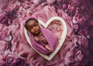 Newborn baby wrapped in purple swaddle sleeping in white heart-shaped bowl surrounded by purple floral fabric.