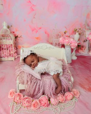 Newborn baby in pink lace dress sleeping on a miniature white bed with roses and pearl accents.