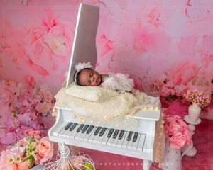 Newborn baby sleeping peacefully on a white toy piano surrounded by pink roses and floral backdrop.