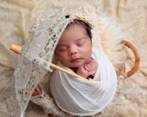 Peacefully sleeping newborn baby wrapped in white swaddle holding a lace parasol in a wicker basket.