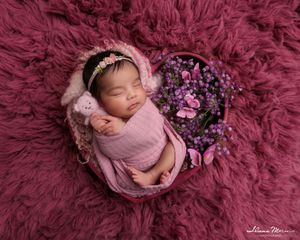 Sleeping newborn baby girl wrapped in pink swaddle inside a heart-shaped box on a mauve floral fur backdrop.