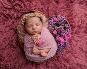 Newborn baby swaddled in pink resting in a wicker basket with purple and pink flowers.