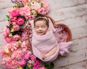 Swaddled newborn baby smiling while resting in a heart-shaped bowl surrounded by pink flowers.