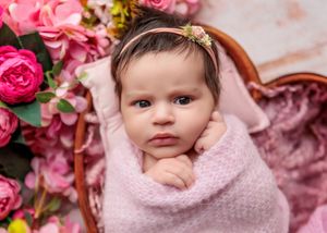 Wide-eyed newborn baby wrapped in pink knit swaddle resting in a basket with pink peony flowers.