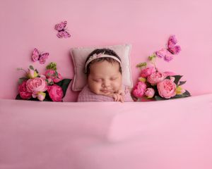 Newborn baby wrapped in pink sleeping on a pink bed surrounded by peonies and butterflies.