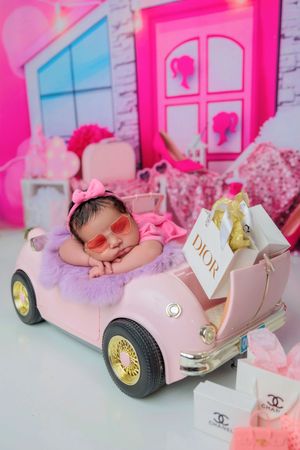 Newborn baby in pink outfit and sunglasses posed in a pink toy car with Dior and Chanel props.