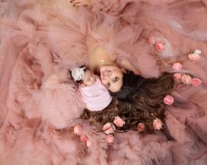 Mother in pink tulle gown lying with her newborn baby surrounded by soft pink roses overhead view.