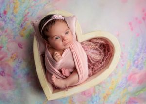 Awake newborn baby girl wrapped in pink swaddle lying in a heart-shaped wooden bowl prop.