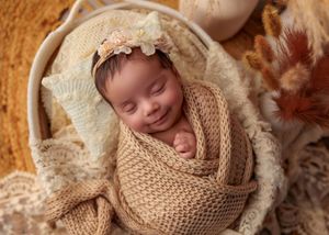 Smiling sleeping newborn baby wrapped in beige knit swaddle in a basket with boho floral decor.