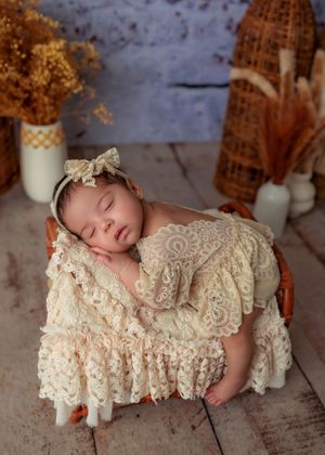Peaceful sleeping newborn in cream lace outfit resting in a wicker basket with dried floral backdrop.