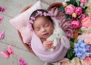 Sleeping newborn in pink swaddle holding white bunny in basket surrounded by colorful spring flowers.