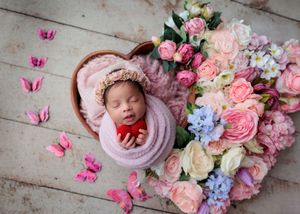Newborn baby wrapped in pink swaddle sleeping in heart-shaped bowl surrounded by colorful spring flowers and butterflies.