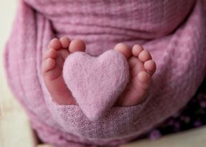 Close-up of newborn tiny feet holding a pink felt heart wrapped in soft purple fabric.